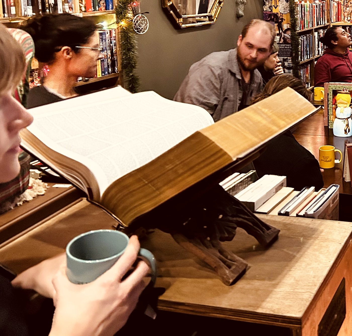 A full-house sitting: readers packed around the long tables of a lamplit bookshop, a huge open book and a mug of tea in the foreground